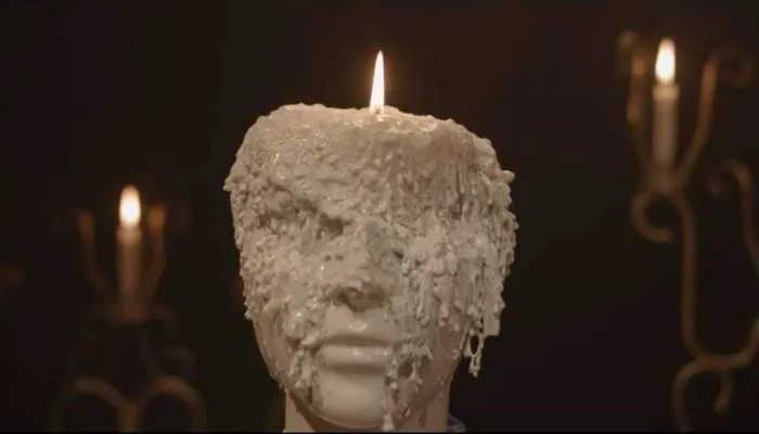 Busto de cera derretida con una vela encendida sobre la cabeza, entre candelabros