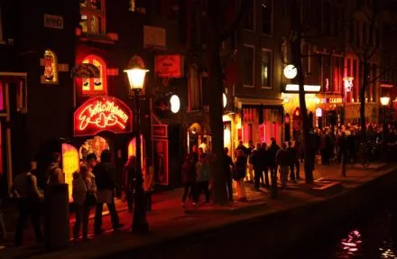 Calle junto a un canal de noche con letreros de neón rojos y transeúntes, en un barrio de vida nocturna
