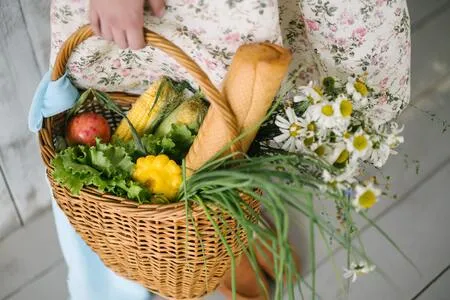 Canasta de mimbre con pan, verduras y flores de manzanilla sostenida por una persona con vestido floreado