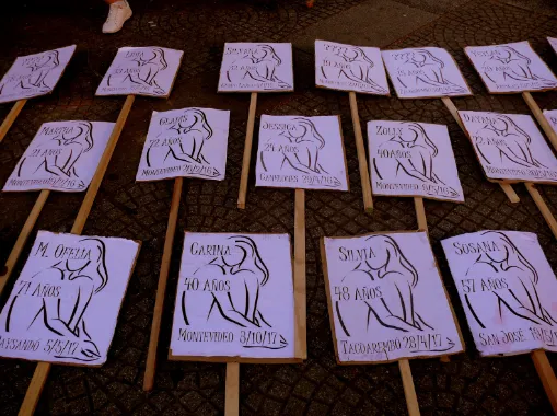 Hileras de carteles blancos con siluetas dibujadas, nombres y edades de mujeres víctimas de feminicidio, sobre el pavimento