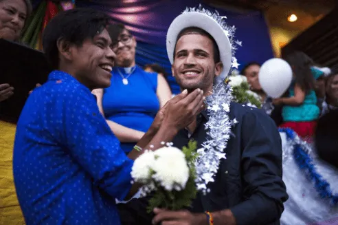 Dos hombres sonríen en una celebración; uno lleva sombrero blanco con adornos plateados y un ramo de flores blancas