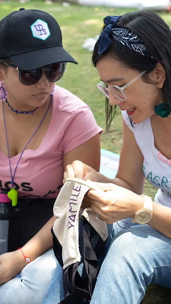 Dos mujeres con gorra y gafas de sol bordan juntas una tela durante el 8M en Bucaramanga