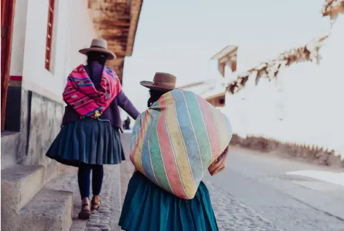 Dos mujeres de espaldas con sombreros, faldas y mantas de colores cargadas caminan por una calle