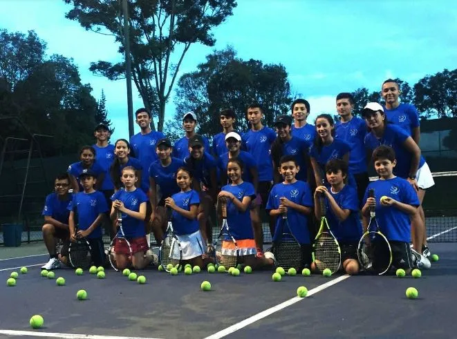 Grupo de niñas y niños con camisetas azules posa con raquetas en una cancha de tenis, rodeados de pelotas