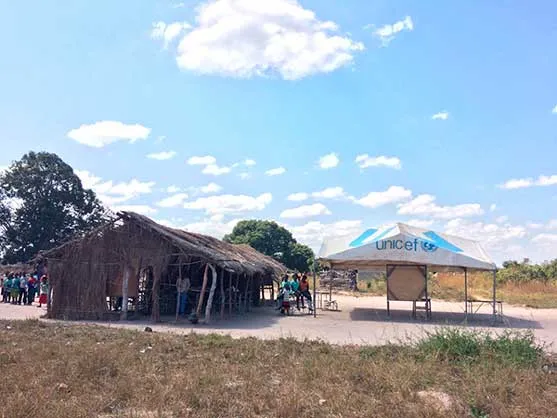 Escuela comunitaria de Mbala con techos de paja y una carpa de Unicef bajo un cielo con nubes, en Zambia