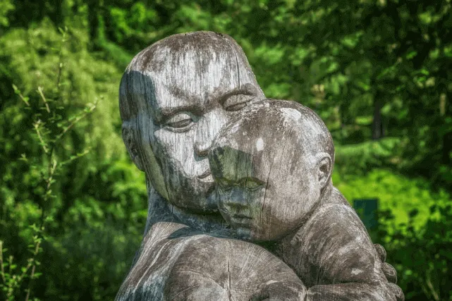 Escultura de piedra de un adulto que abraza a un niño, entre vegetación verde