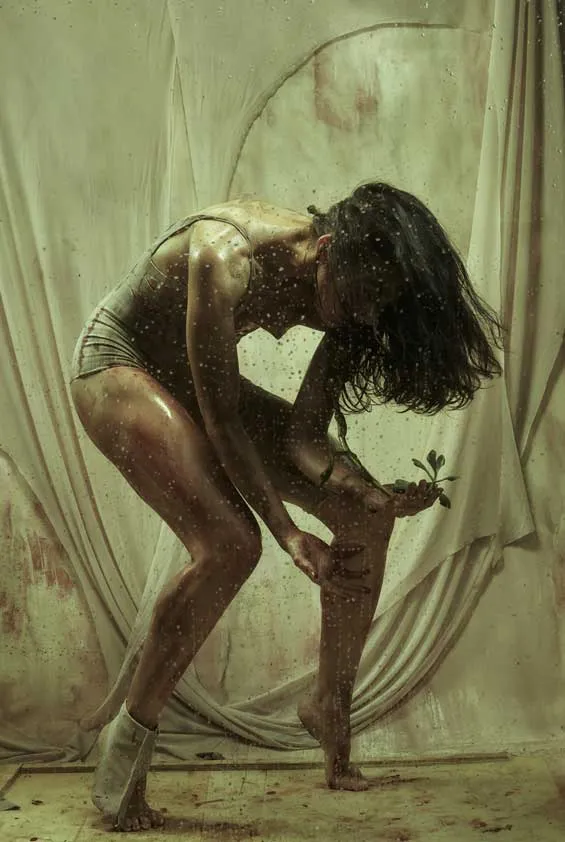 Bailarina agachada con el cabello sobre el rostro sostiene una pequeña planta ante una tela vieja