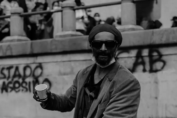 Hombre con boina y gafas de sol sostiene un vaso frente a un muro con la pinta «Estado asesino», en blanco y negro