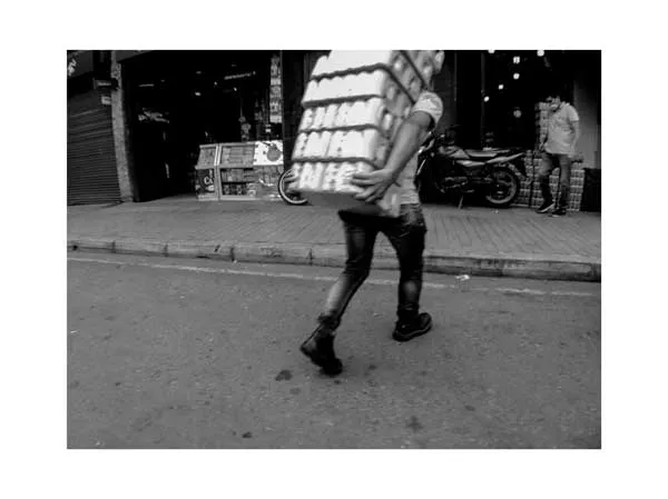 Hombre camina por la calle cargando en el hombro una pila de cartones de huevos, en blanco y negro