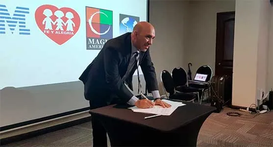Hombre de traje firma un documento sobre una mesa frente a una pantalla con los logos del acuerdo