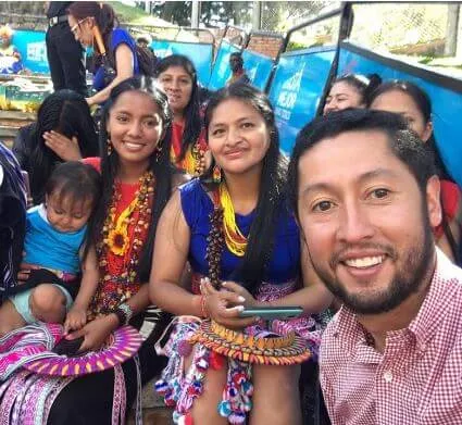 Hombre sonriente se toma un selfi con mujeres indígenas con trajes tradicionales de colores, una con un bebé
