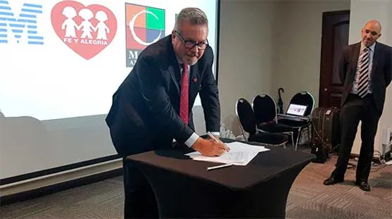 Hombre de traje firma un documento en la mesa mientras otro hombre lo observa de pie