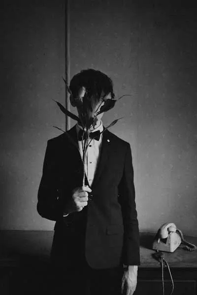 Hombre de traje y moño en blanco y negro con una flor cubriéndole el rostro, junto a un teléfono antiguo