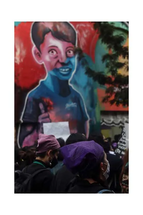 Mural de un niño de rostro azul en una pared, con manifestantes de gorro morado debajo