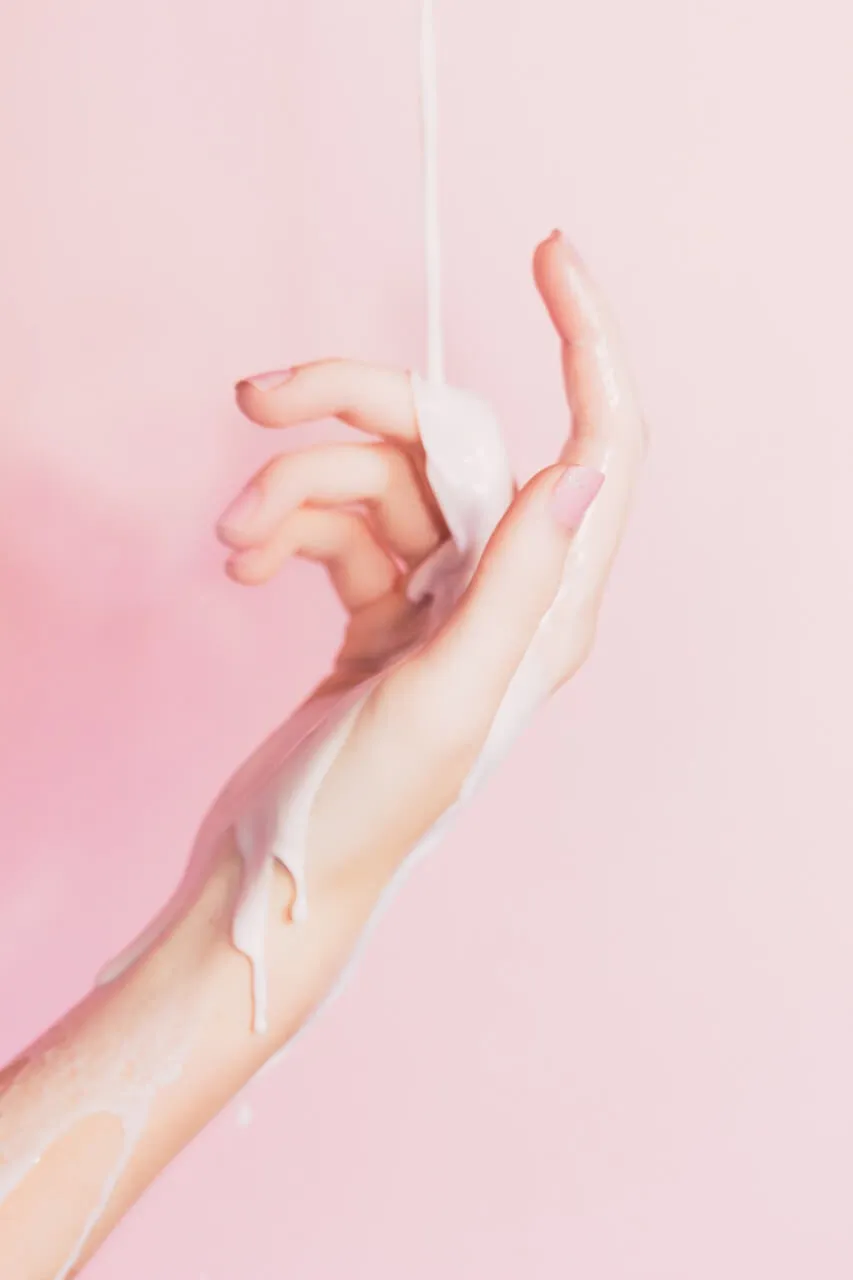 Ilustración de una mano que sostiene un tampón por el hilo, con líquido blanco que escurre, sobre fondo rosa