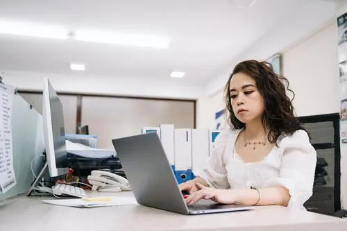 Joven de blusa blanca escribe en una laptop en una oficina con archivadores y escritorios
