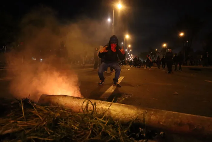 Manifestante agachado tras un tronco caído junto a un fuego en la calle, de noche