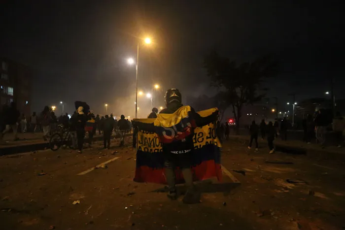 Manifestante envuelto en una bandera de Colombia de pie en una calle oscura, con otros manifestantes al fondo
