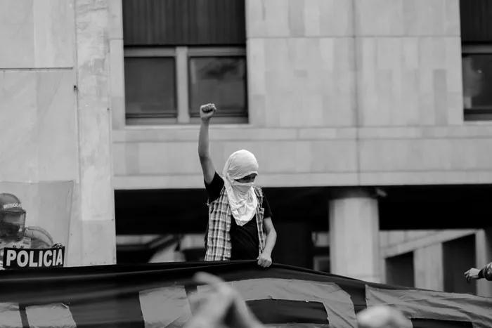 Manifestante con el rostro cubierto levanta el puño desde una estructura frente a un edificio con letrero de policía