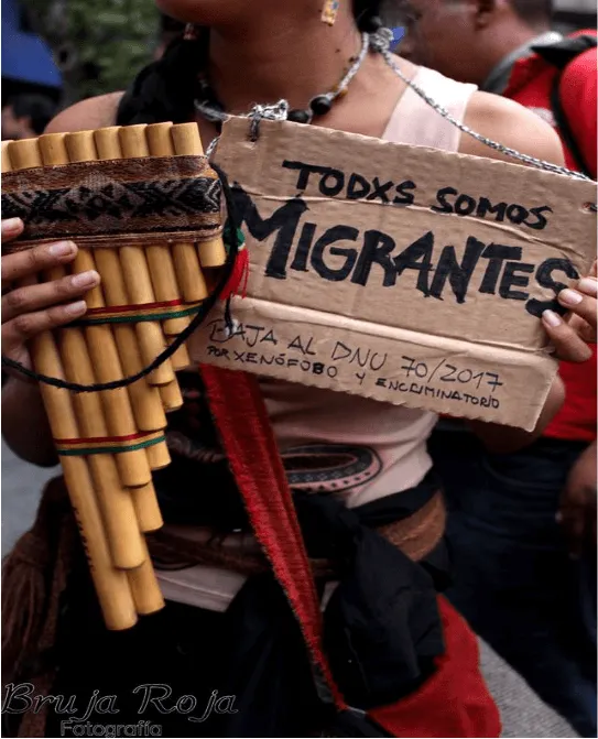 Manifestante con una zampoña sostiene un cartel de cartón «Todxs somos migrantes» en una marcha