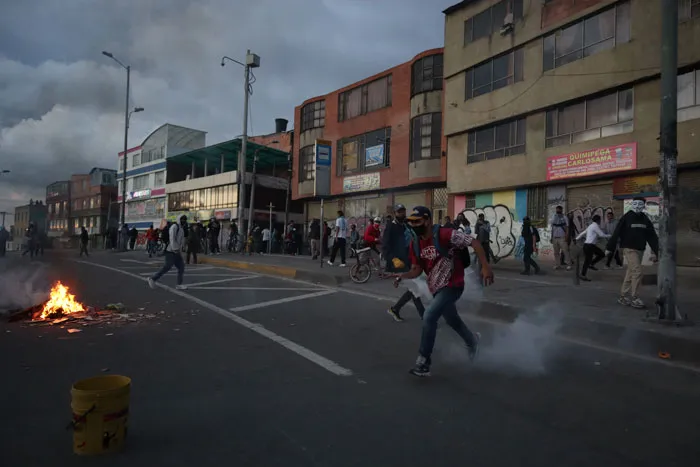 Manifestantes corren por una avenida entre humo, con una fogata al costado y edificios al fondo