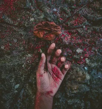 Mano manchada de rojo asoma sobre una superficie de piedra oscura junto a una flor marchita