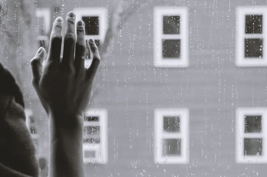 Mano apoyada en el vidrio de una ventana con un edificio de ventanas al fondo, en blanco y negro