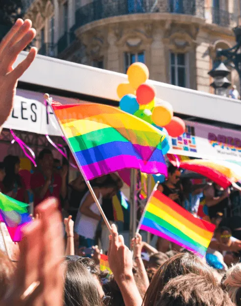 Multitud en una marcha alza banderas del arcoíris y globos de colores frente a edificios de una ciudad