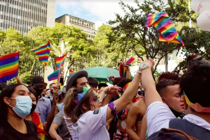 Asistentes con banderas arcoíris y manos en alto bailan entre árboles durante la Marcha del Orgullo en Bogotá