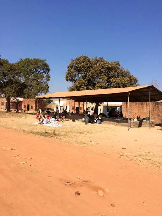 Personas reunidas bajo un techo en un mercado de tierra roja con árboles al fondo, en Chibombo, Zambia