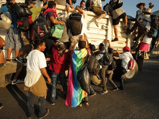 Multitud de migrantes con mochilas sube a un camión, con una persona envuelta en una bandera arcoíris