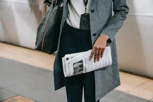Mujer con abrigo gris y bolso sostiene un periódico doblado en una estación