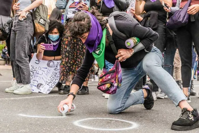 Mujer agachada rocía pintura en aerosol sobre el pavimento, rodeada de manifestantes con pañuelos morados