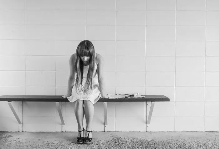 Mujer sentada en una banca con la cabeza baja frente a un muro blanco, en blanco y negro