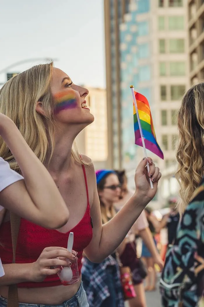 Mujer con la cara pintada de arcoíris sostiene una pequeña bandera LGBT+ entre la gente en una ciudad