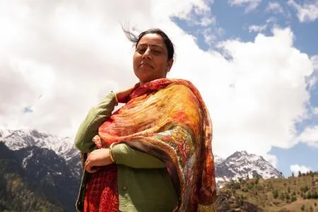 Mujer con chal de colores naranja y rojo posa de pie frente a montañas nevadas y un cielo con nubes