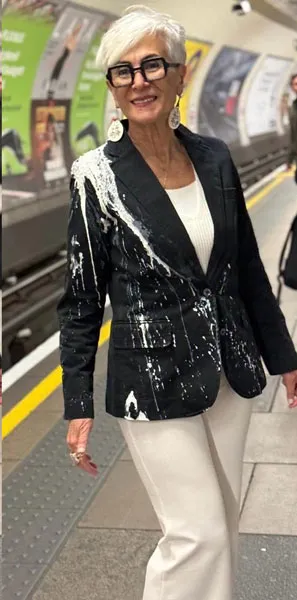 Mujer mayor de cabello canoso y gafas con chaqueta negra restaurada con detalles blancos, de pie en una estación de metro