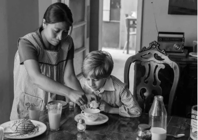Mujer con delantal sirve el desayuno a un niño sentado a la mesa, en blanco y negro