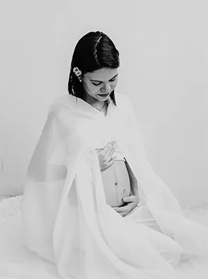 Mujer embarazada envuelta en una tela blanca, sentada con la mirada baja y las manos sobre su vientre, en blanco y negro