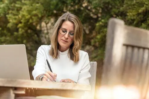 Mujer con gafas y blusa blanca escribe en una libreta junto a una laptop en una mesa al aire libre