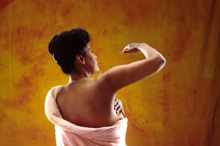 Mujer de espaldas se palpa el brazo y la axila en un autoexamen de mama, con un albornoz rosado