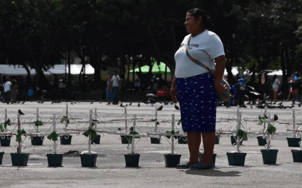 Mujer de pie frente a hileras de cruces con plantas en una plaza, en memoria de las niñas de Guatemala