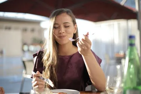 Mujer joven de cabello largo come con tenedor en un restaurante, con los ojos cerrados