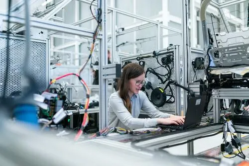 Mujer con gafas trabaja en una laptop junto a maquinaria de un laboratorio de ingeniería