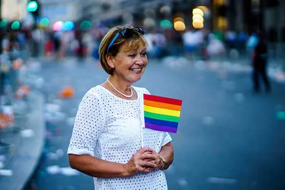 Mujer mayor sonriente sostiene una pequeña bandera arcoíris en un desfile del orgullo