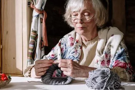 Mujer mayor con gafas y chal floreado teje con lana gris sentada a una mesa