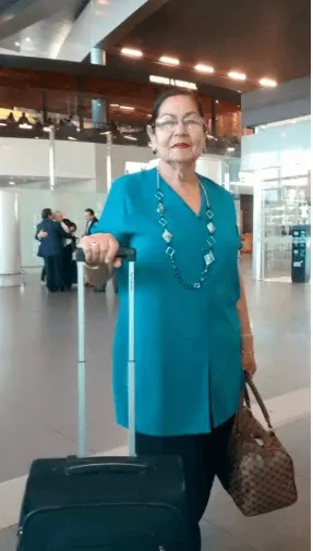 Mujer mayor con blusa turquesa sostiene una maleta de ruedas y un bolso en un aeropuerto