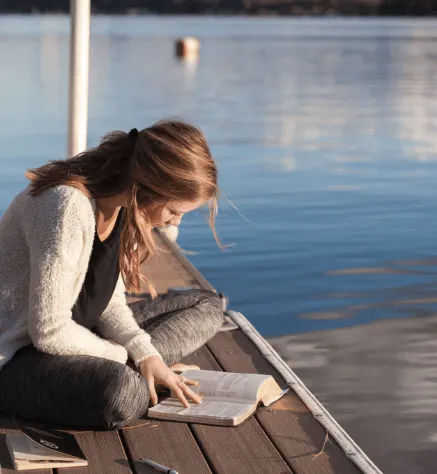 Mujer sentada en un muelle junto a un lago lee un libro abierto sobre sus rodillas
