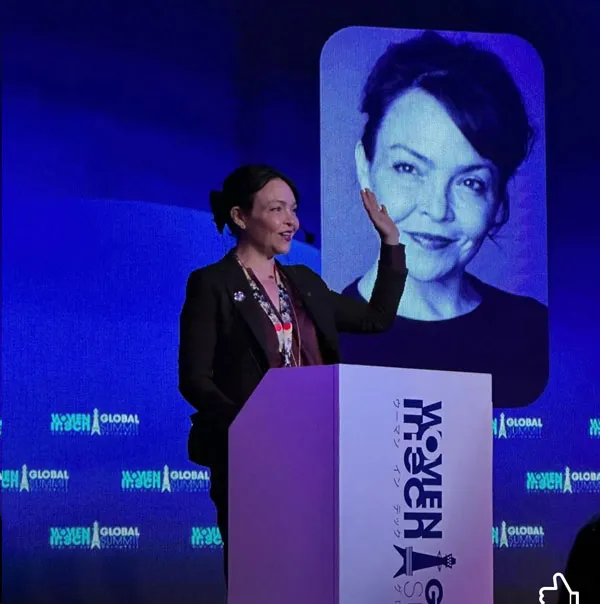 Mujer habla desde un podio de Women in Tech Global, con el retrato de una mujer proyectado en la pantalla detrás