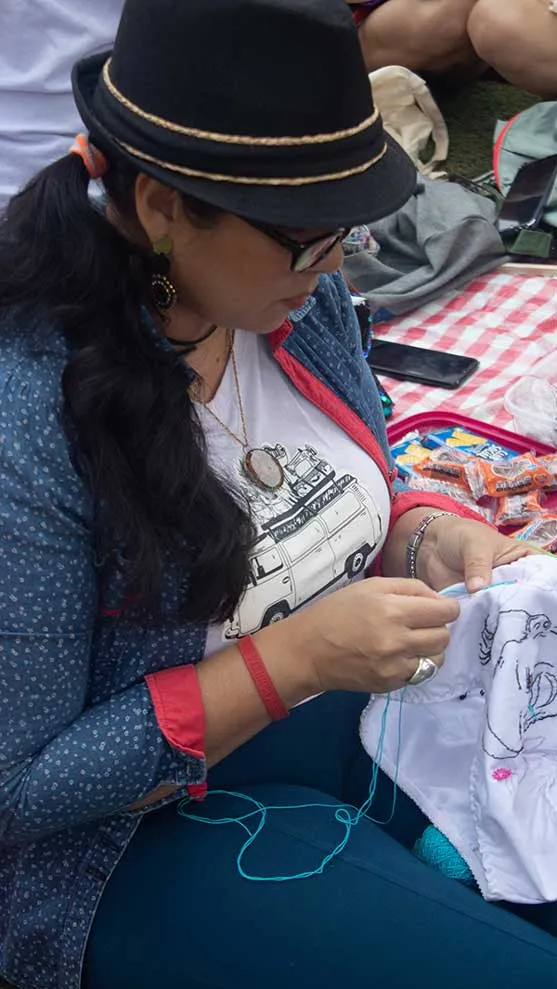 Mujer con sombrero y chaqueta azul bordando una tela blanca en un parque